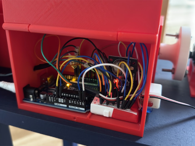 The red 3D-printed electronics box with its lid open, showing the Arduino, real-time clock module and wiring inside, mounted under the hopper on the painted frame.