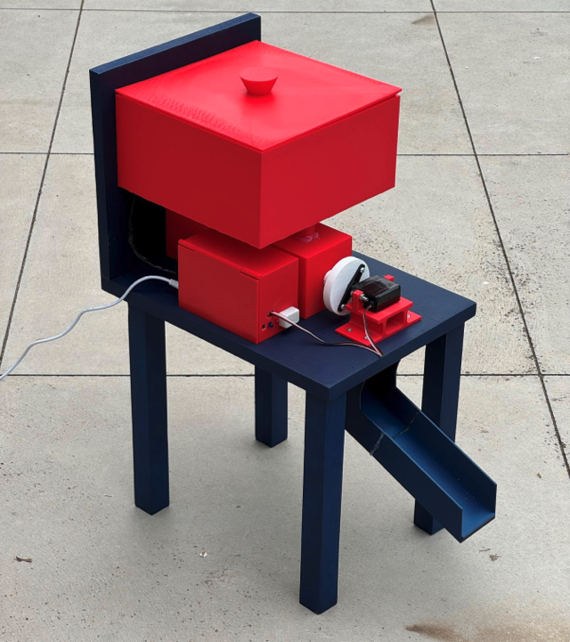 The finished dog feeder: a red 3D-printed hopper and electronics box mounted on a navy-painted timber frame, with a chute running down to a dog bowl.